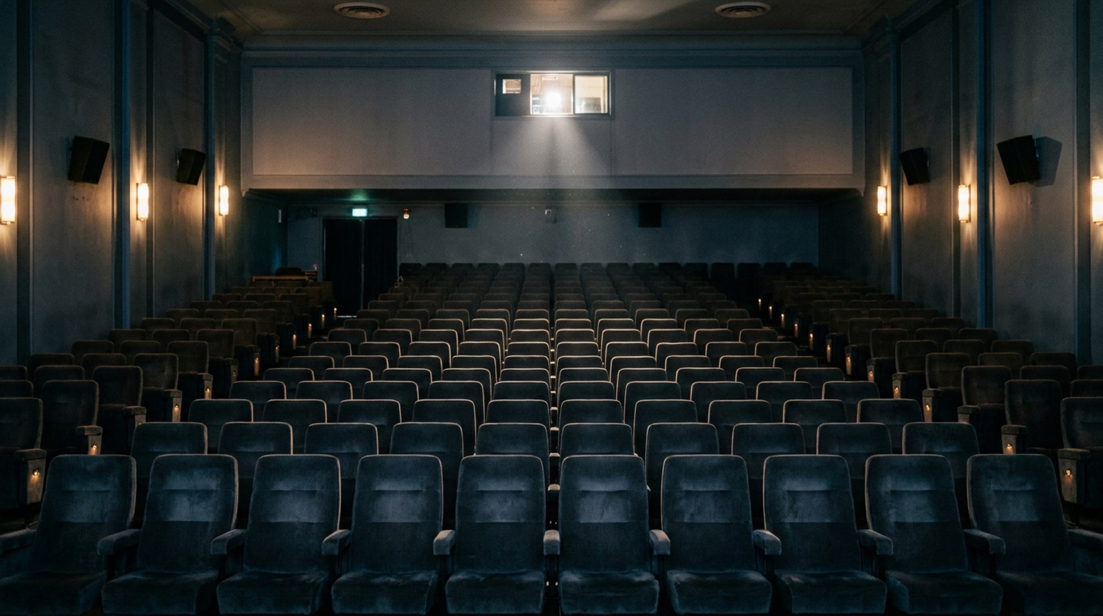 Gösterimden önce ışıkları henüz sönmemiş, koltukları boş bir sinema salonunun perde tarafından bakışı; perde grisi ve loş duvar lambaları.