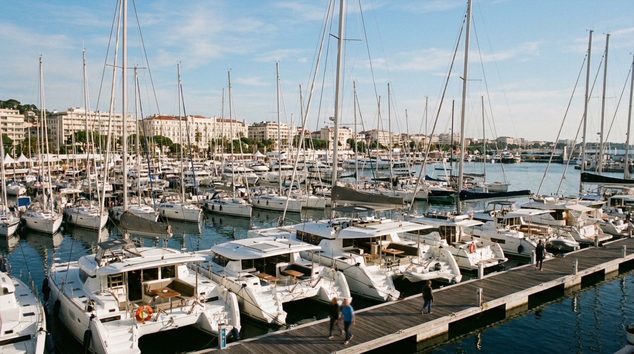 Cannes Yachting Festival'de liman dolu: yelkenliler ve katamaranlar pruva iskeleye yanaşmış, Croisette binalar fon