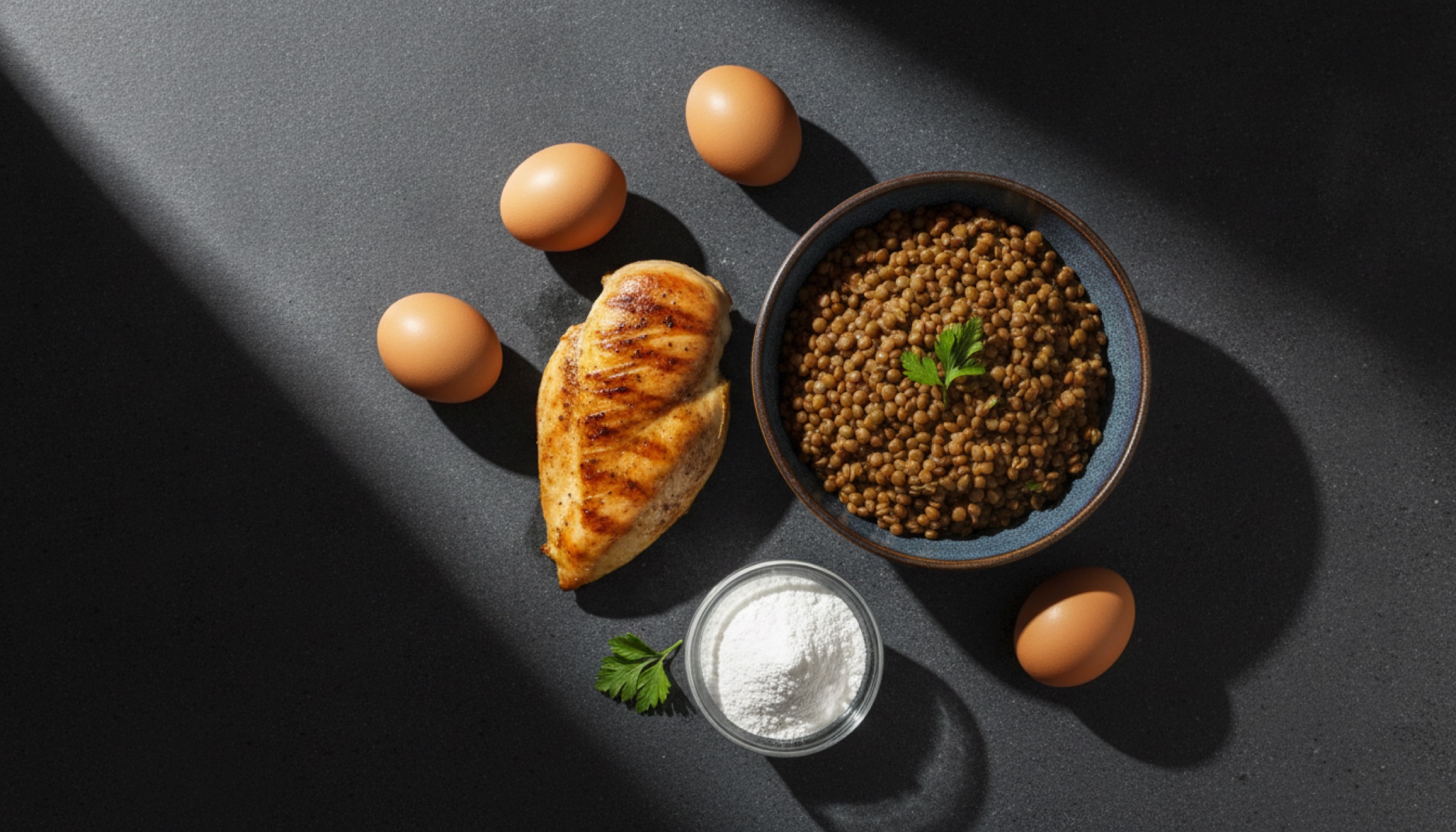 Koyu taş yüzey üzerinde yumurta, tavuk, mercimek ve protein tozu — dramatik yan aydınlatma ile üstten çekim