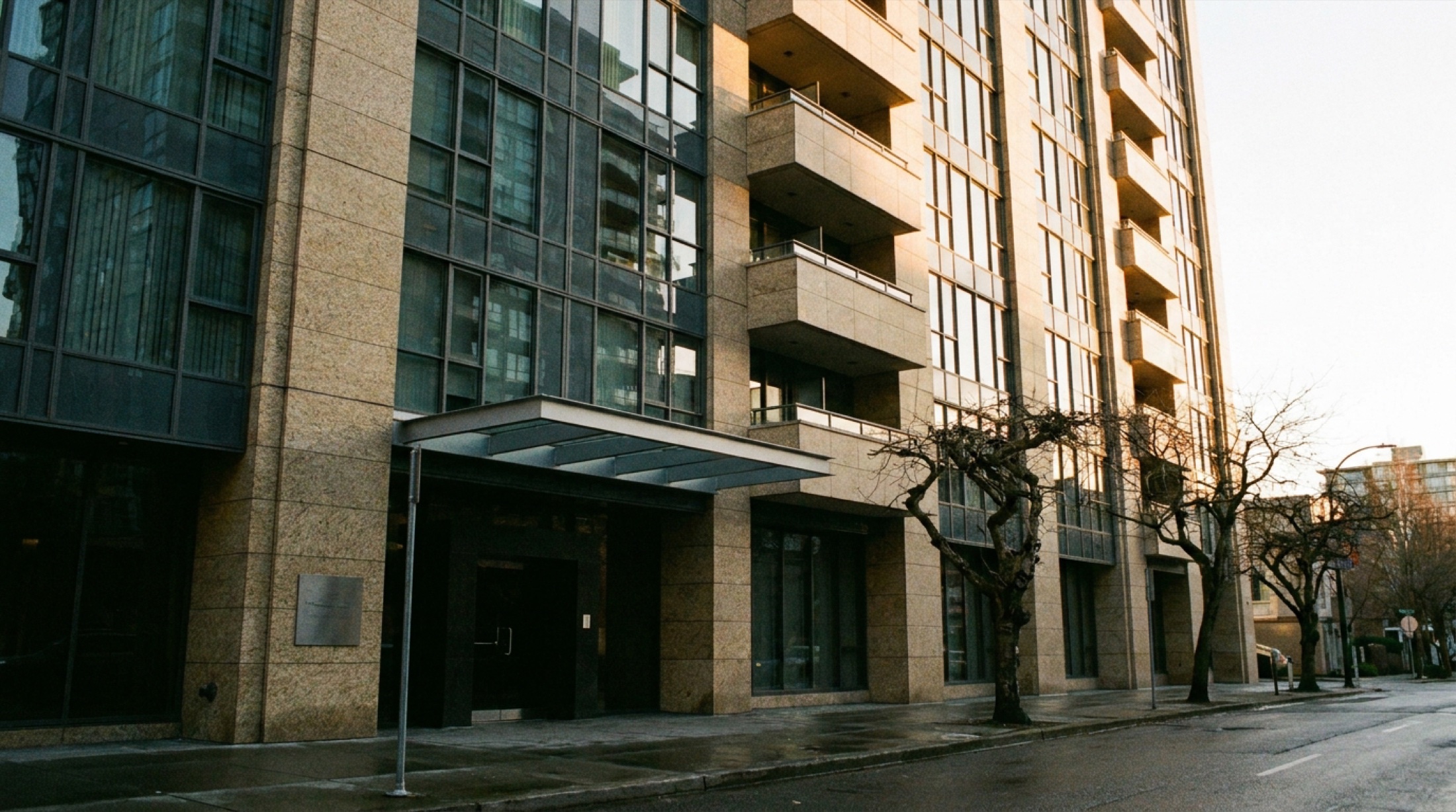 Büyük bir şehirde modern bir konut kulesinin sokak seviyesinden yukarı bakılarak çekilmiş cephesi; cam ve taş kaplama, giriş sundurmasında okunmayan metal bir tabela, öğleden sonra ışığı.