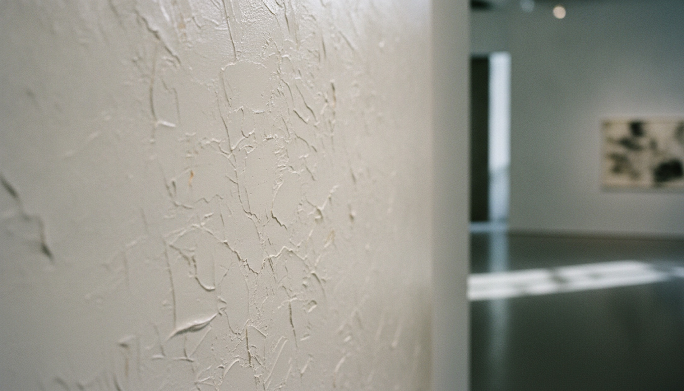 Barbican galerisinde beyaz boyalı tuval önünde, yan ışığın yüzey dokusunu belirginleştirdiği bir kurulum