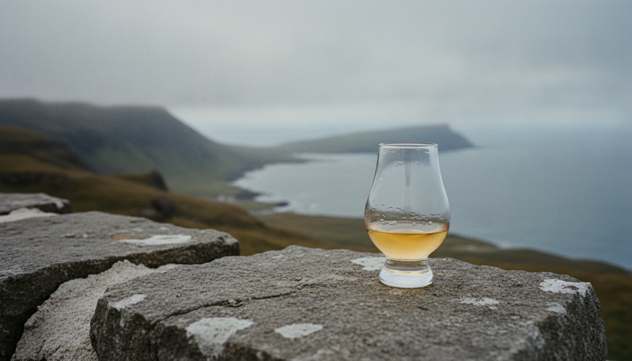 Skye sahilinin sisli kayalıkları önünde Glencairn kadehinde soluk altın renkli Torabhaig Taigh single malt viskisi