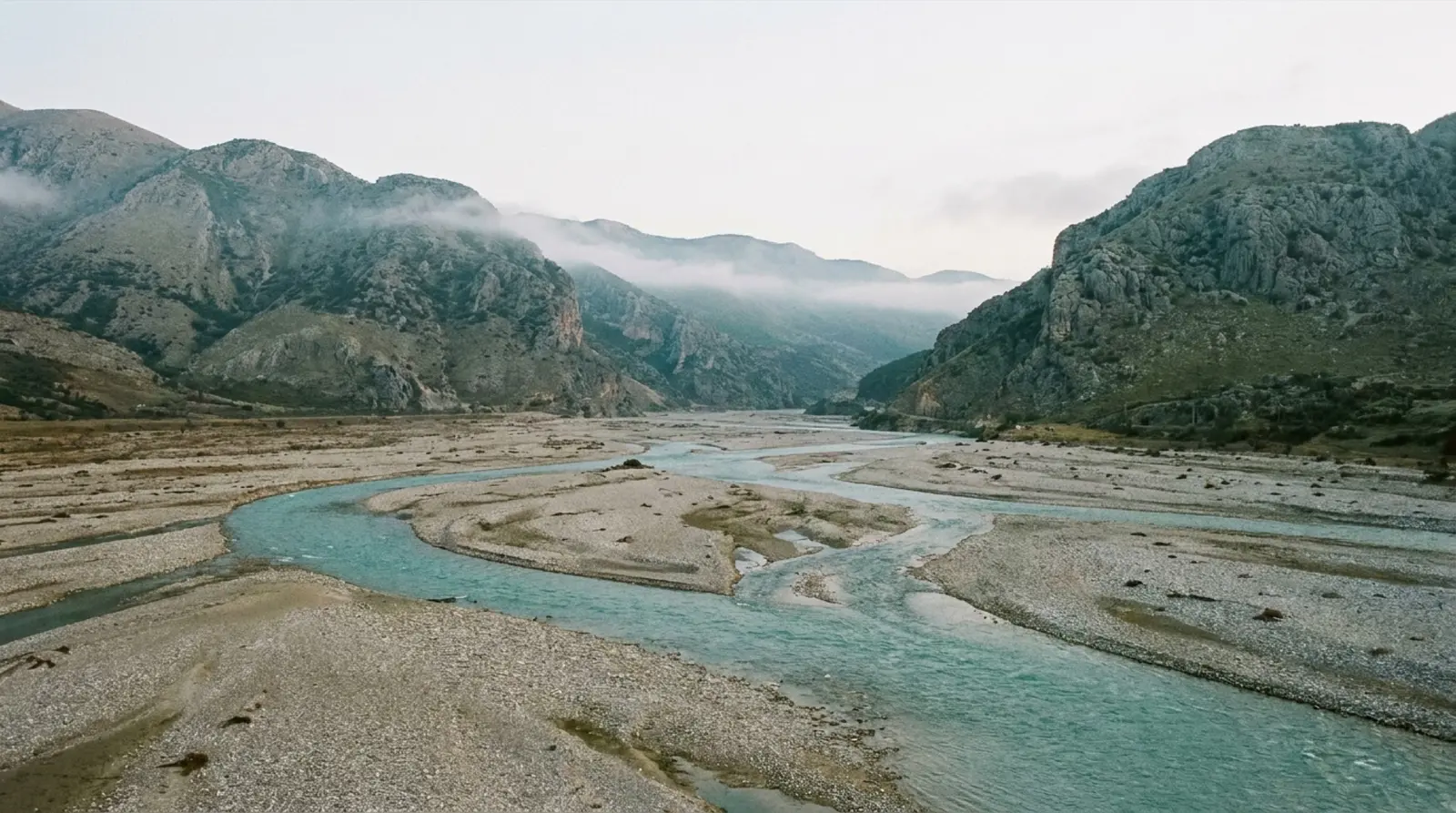 Arnavutluk'ta Vjosa Nehri'nin örgülü kollara ayrılmış geniş çakıl yatağı, arkada Tepelenë çevresindeki çıplak dağlar, insan yok