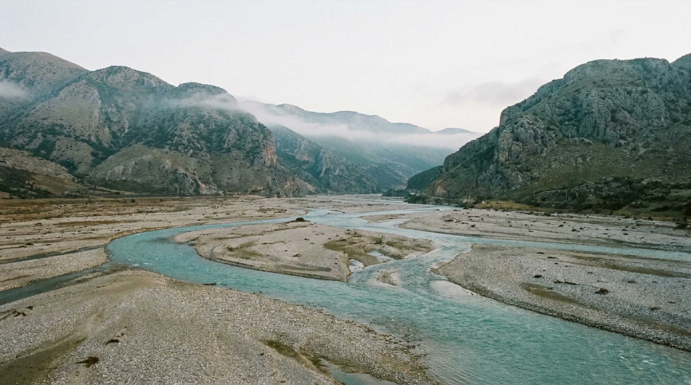 Arnavutluk'ta Vjosa Nehri'nin örgülü kollara ayrılmış geniş çakıl yatağı, arkada Tepelenë çevresindeki çıplak dağlar, insan yok